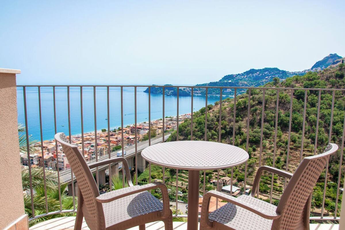 A balcony with wicker chairs and a round table, overlooking a coastal town and the sea.
