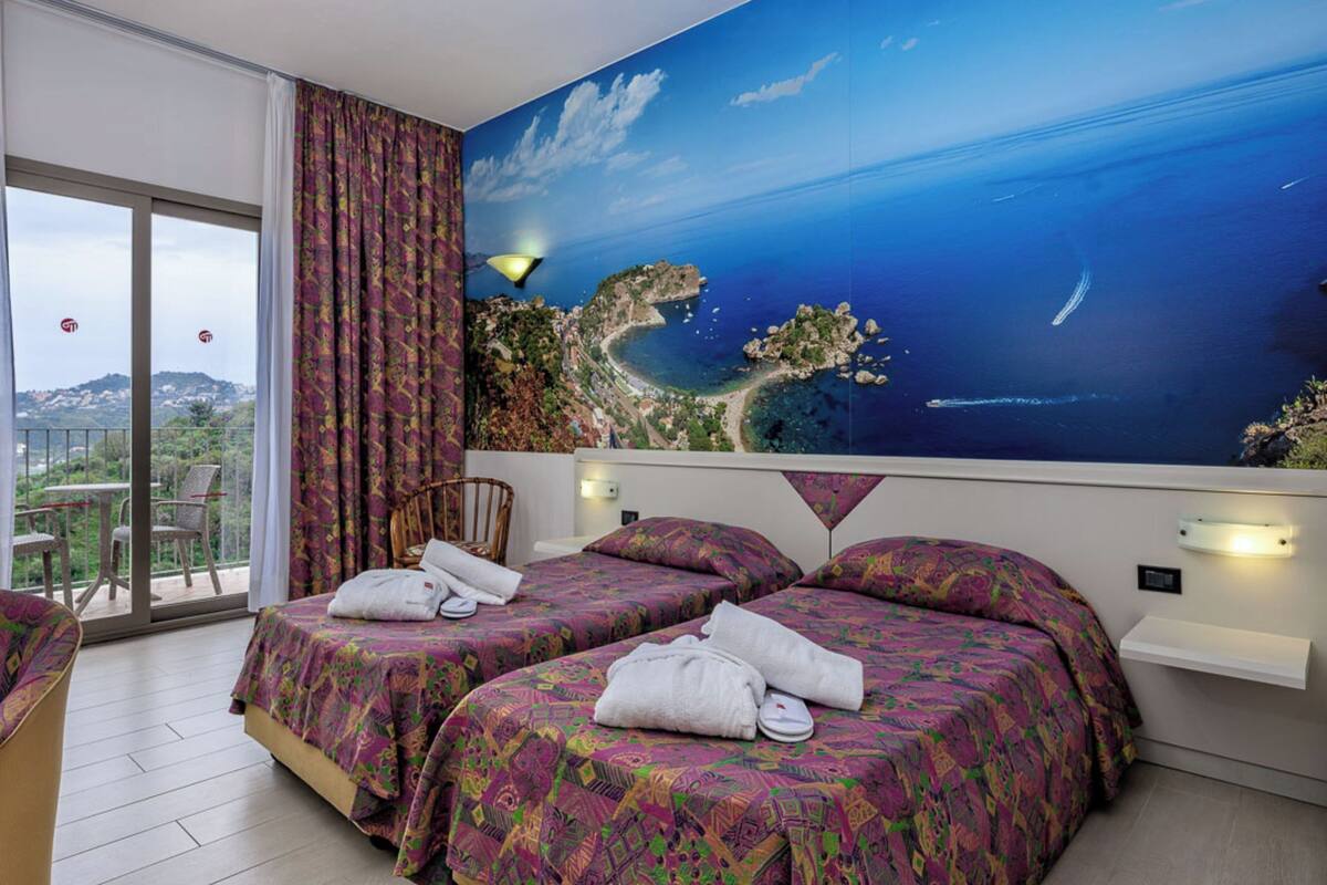 A hotel room with two beds, a balcony with a view of the sea, and a large mural of a coastal landscape.