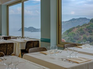 A restaurant with tables set for dining, large windows offering a view of a coastal landscape. - Antares (Letojanni)