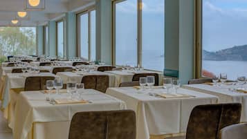 Un restaurant doté de nombreuses tables dressées pour le service, de grandes fenêtres offrant une vue sur la mer et d’un décor intérieur moderne.