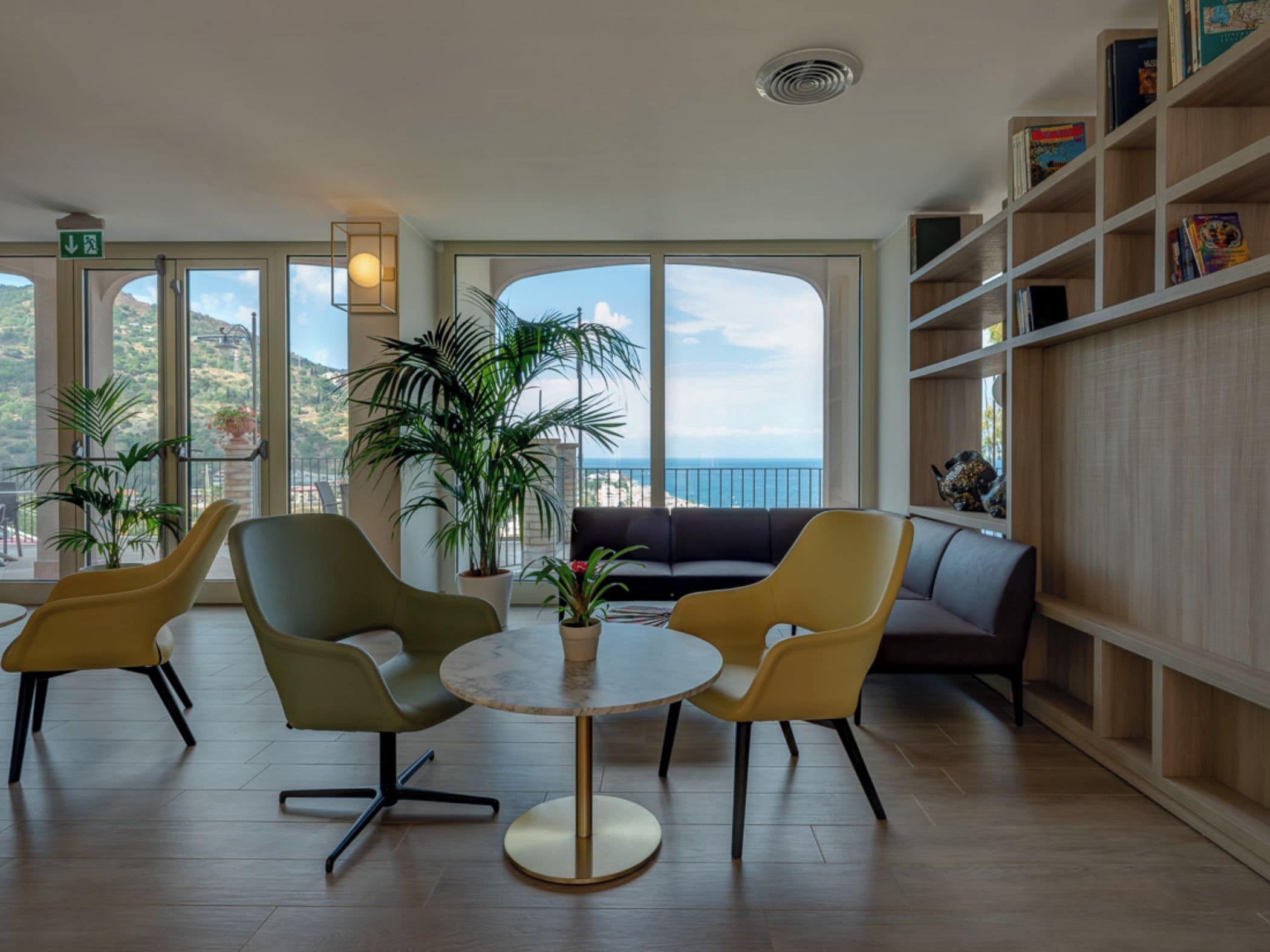 Un salon moderne avec une table ronde et des chaises, un canapé et une bibliothèque. De grandes fenêtres offrent une vue sur la mer et les collines.