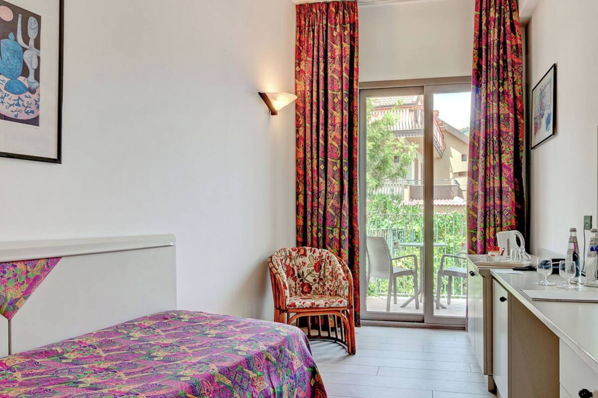 A room with a bed, a chair, a desk with a chair, and a balcony with a view.
