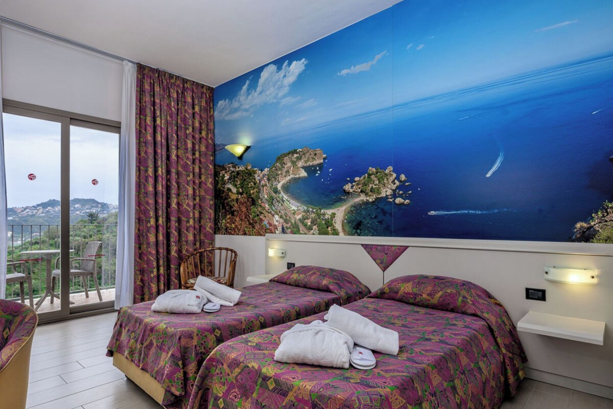 Habitación de hotel con dos camas, un balcón con vista al mar y un mural grande de un paisaje costero.