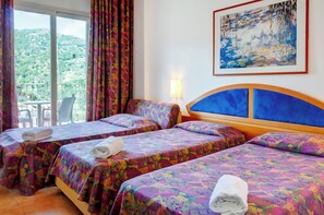 Ein Hotelzimmer mit zwei Betten, farbenfrohen gemusterten Bettdecken, einem Bild an der Wand und einem Balkon mit Blick auf Grünflächen.