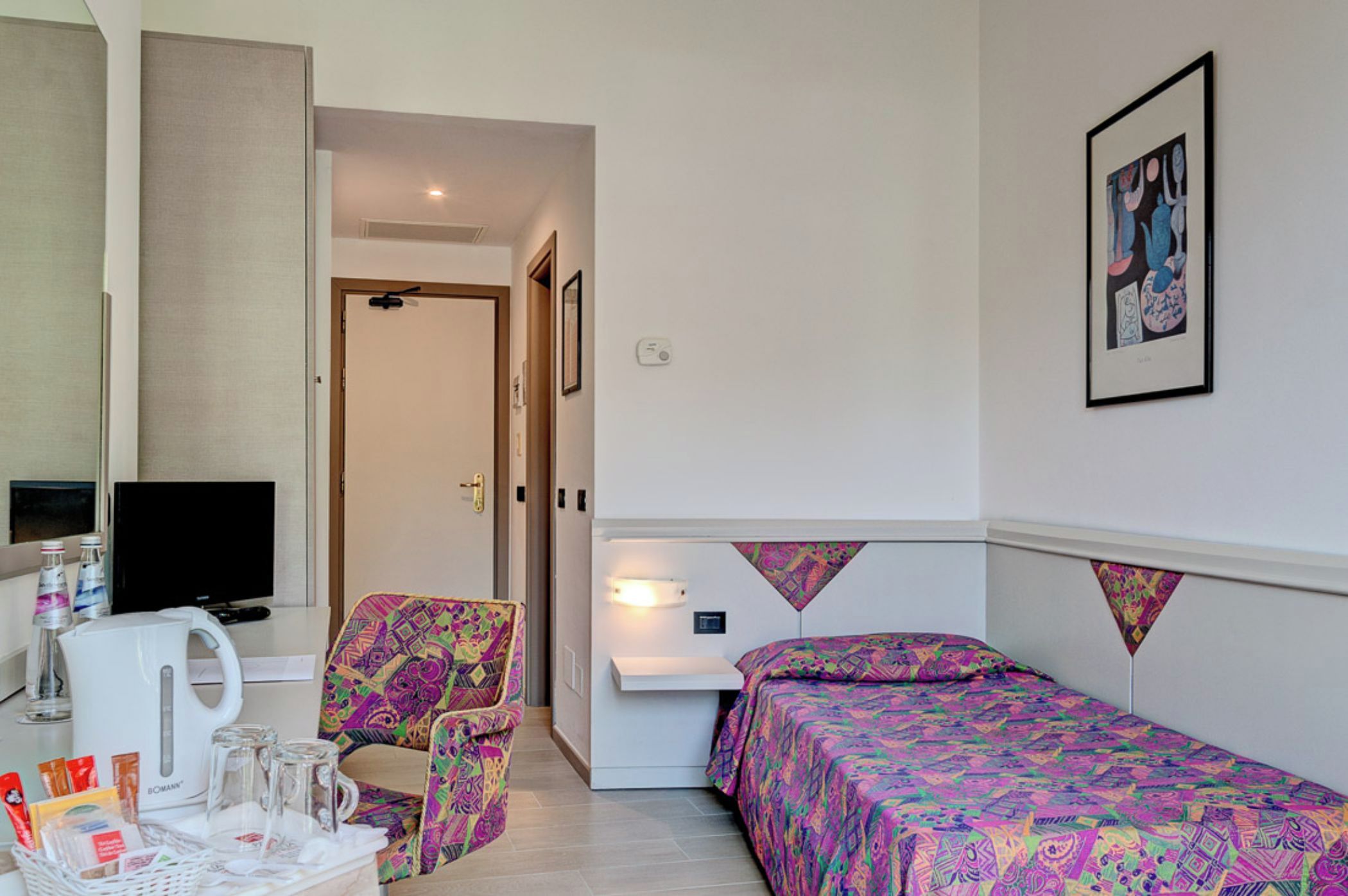 A hotel room with a bed, a chair, a desk with a kettle, a television, and a framed picture on the wall.