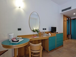 Une chambre d’hôtel compacte comprenant un bureau en bois, une chaise, une petite table ronde, un miroir, un téléviseur et une armoire de couleur turquoise.
