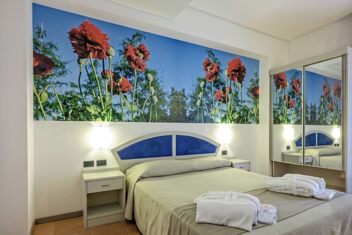 Una habitación de hotel con una cama, una mesita de noche, un espejo y un cuadro grande con motivos florales en la pared.