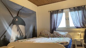 Een slaapkamer met een bed, een raam met gordijnen, een radiator en een muurschildering van een molen.