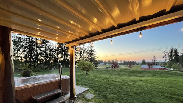 A covered outdoor area with a hot tub, steps leading to it, and a view of a grassy field and trees in the background.