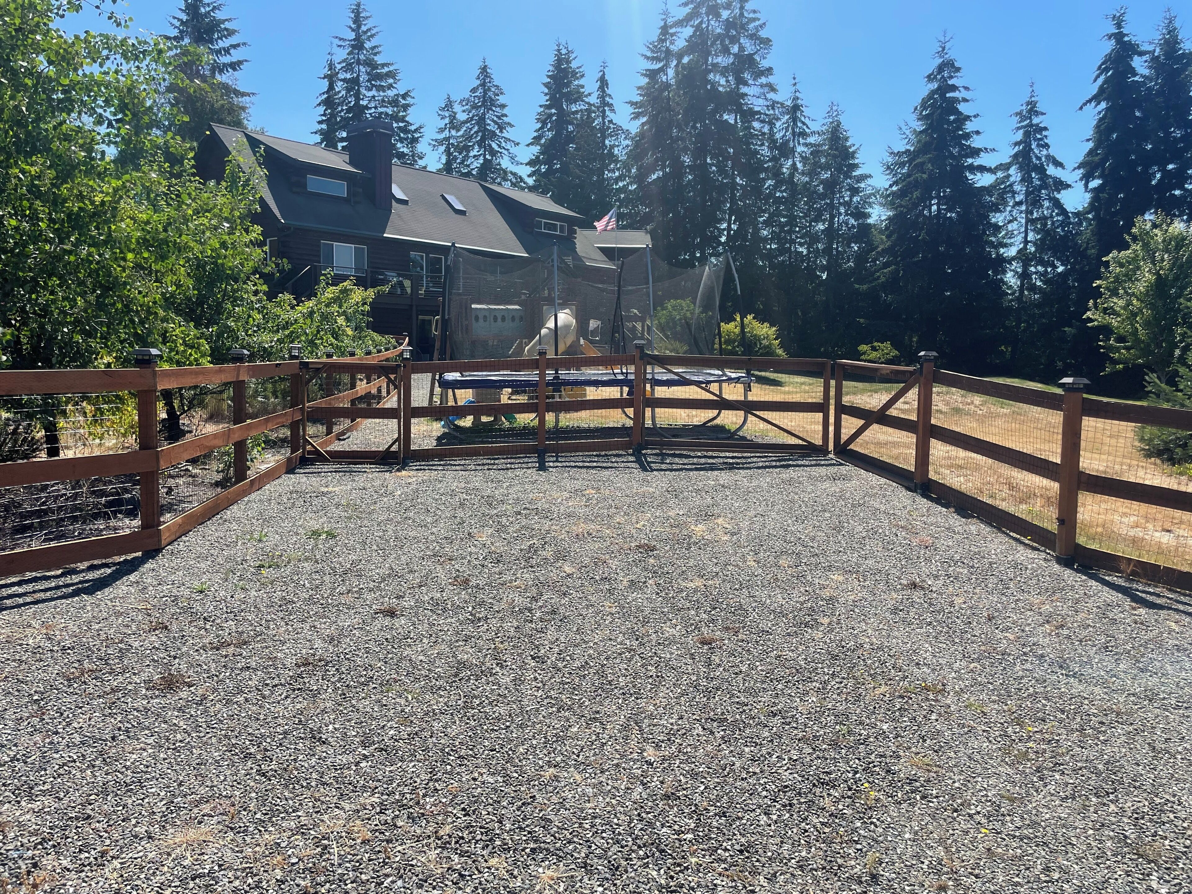 A gravel driveway leading to a wooden gate with a trampoline and a covered play area in the background.
