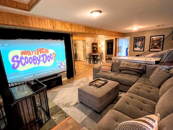 Un soggiorno con una televisione che mostra 'What's New Scooby-Doo?', un divano grigio, un tavolino da caffè e un tavolo da biliardo sullo sfondo.