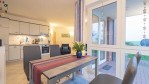 Een moderne keuken met witte kasten, een eethoek met een geruit tafelkleed, en uitzicht op een bakstenen gebouw door grote ramen.