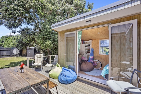 A modern outdoor living space with a wooden deck, seating area, and a glass door leading to a room with a bicycle and bookshelves.