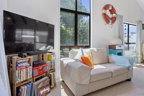 A living room with a flat-screen TV, a bookshelf with vinyl records, a beige sofa, and a large window with a view of trees.