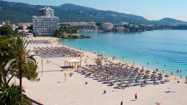 Una platja amb nombrosos para-sols i gandules, un edifici gran al fons, i una vista clara del mar i les colines.