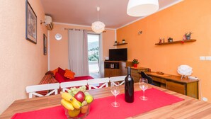 A dining area with a wooden table, red placemat, fruit bowl, wine glasses, and a bottle. A TV and kitchen area are visible in the background. - Apartment Jossy (Dubrovnik)