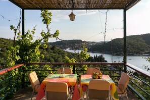 A covered outdoor seating area with tables and chairs, overlooking a scenic landscape with a body of water and hills. - Bodul Vacation Apartment (Dubrovnik)