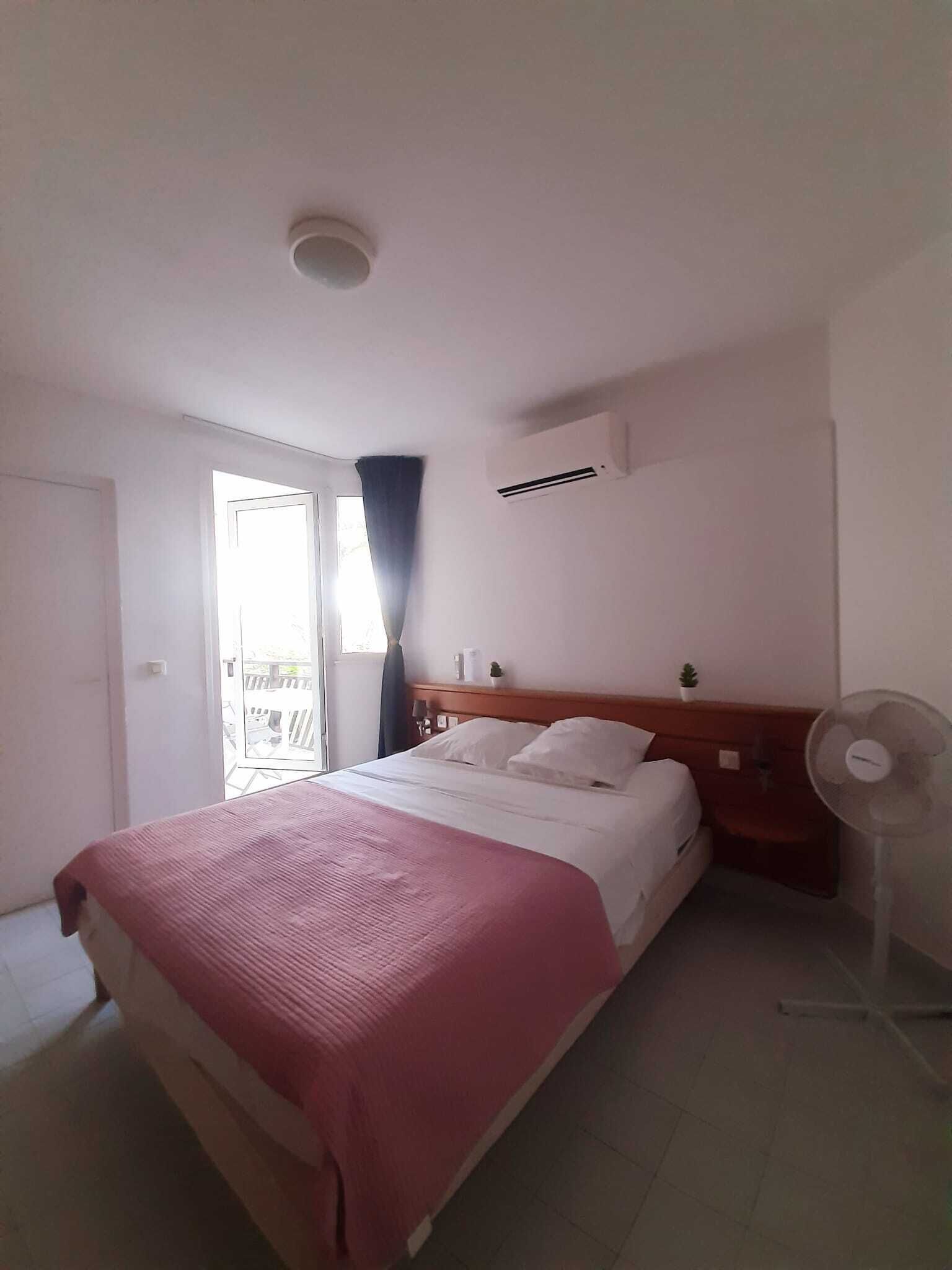 A hotel room with a bed, a window with curtains, an air conditioning unit, and a standing fan.