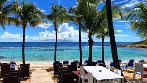 A beachside restaurant with a wooden deck, white tables, and chairs, overlooking the ocean.