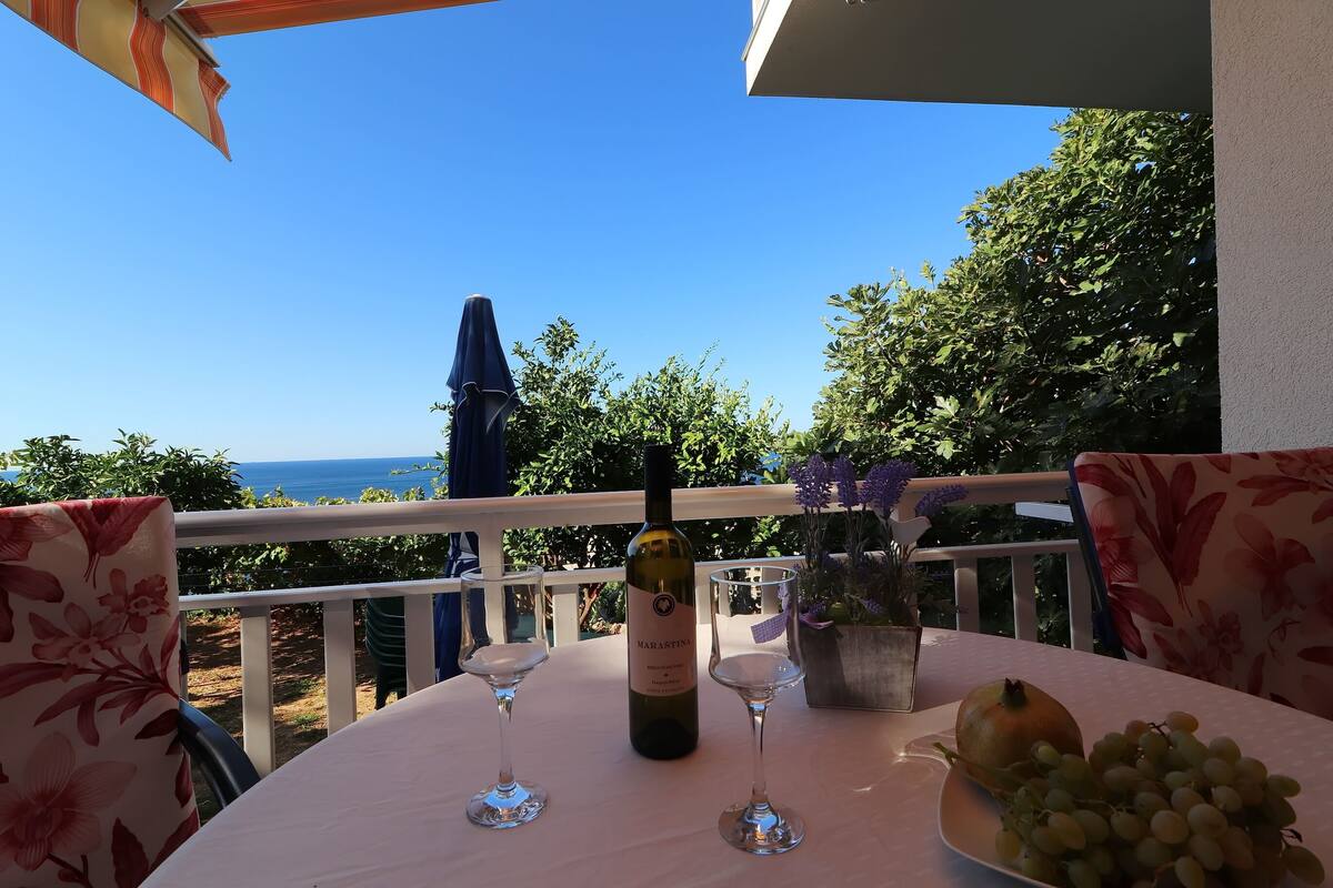 Una mesa con una botella de vino, dos copas de vino y un bol de uvas en un balcón con vistas al mar.