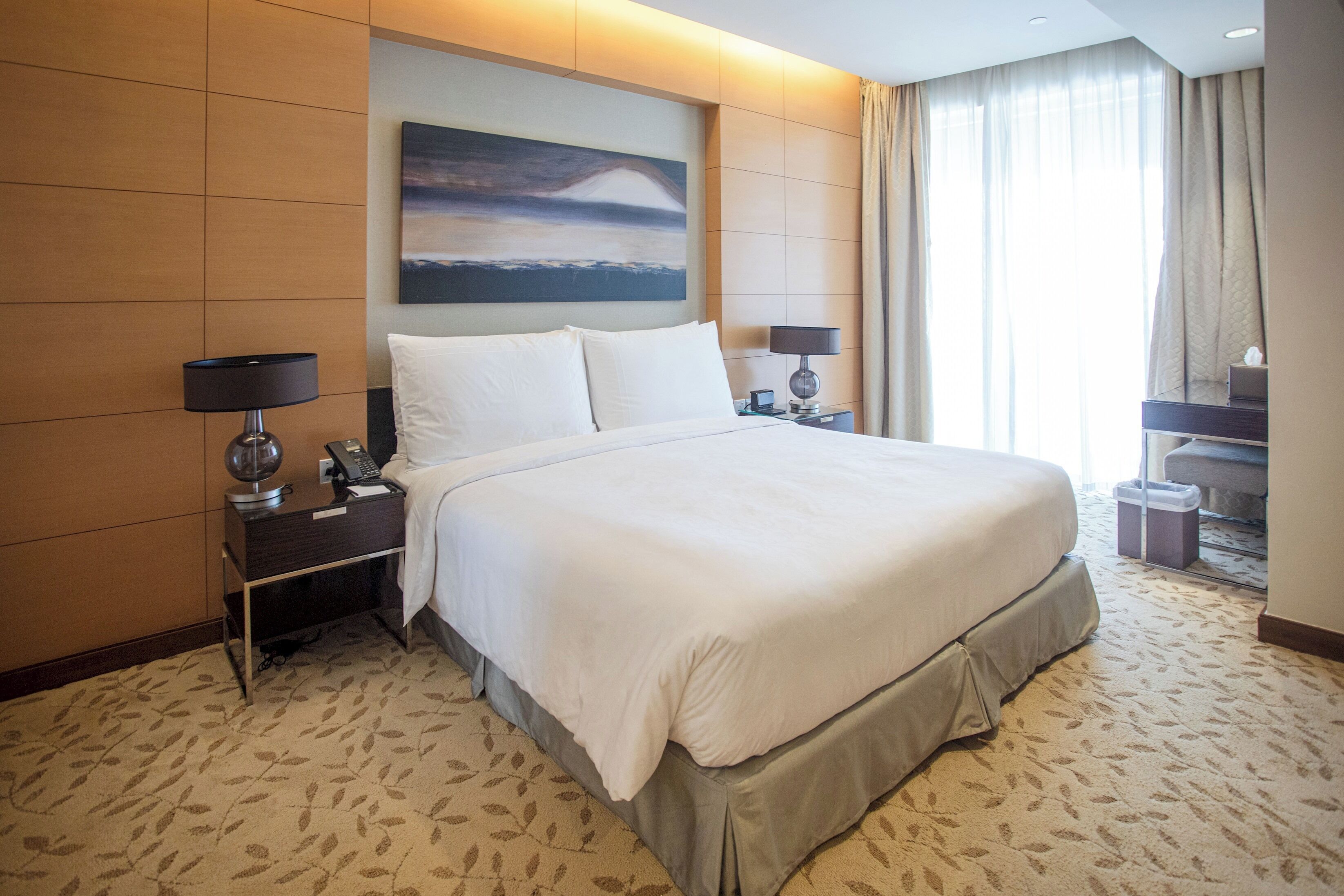 Quarto de hotel com uma cama grande, duas mesas de cabeceira com luminárias, um quadro na parede e vista para o exterior pela janela.