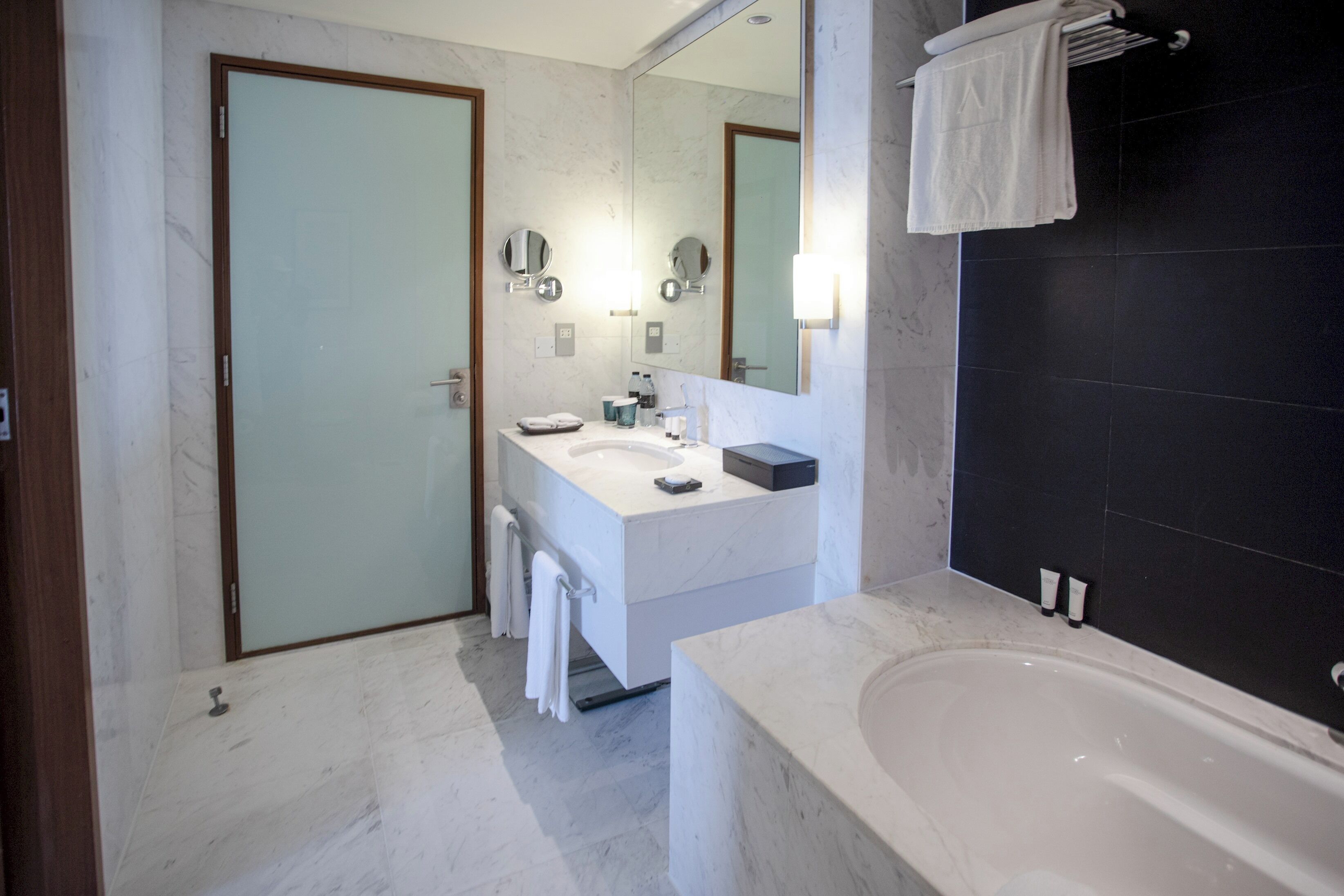 A modern bathroom with a white vanity, a large mirror, a bathtub, and a dark tiled wall.