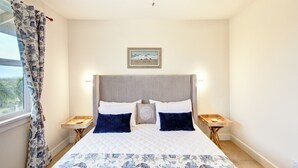 A bedroom with a large bed, two bedside tables, a headboard, a window with curtains, and a wall-mounted picture.