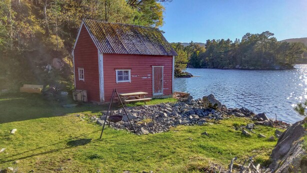 A small red cabin by a lake with a grassy area in front and a rocky shoreline. - Gorgeous home in Reksteren with kitchen (Reksteren)