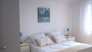 Une chambre à coucher comprenant un lit, deux tables de chevet, une commode, une lampe, un tableau et une fenêtre avec des rideaux.