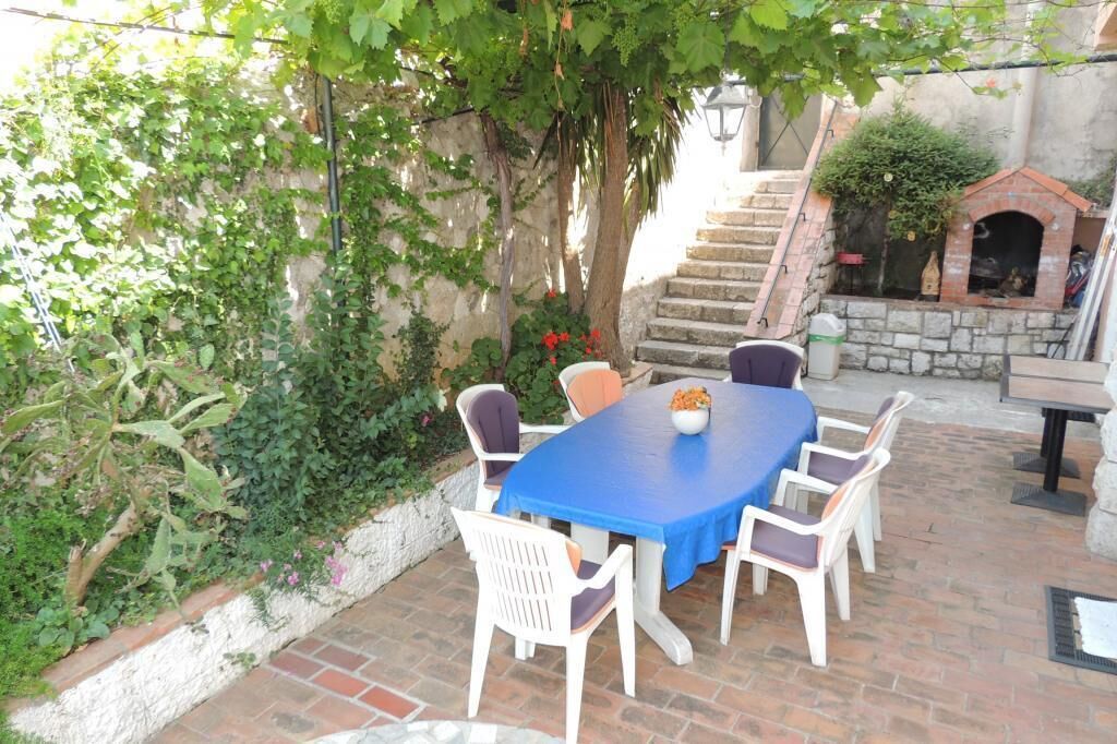 Um pátio externo com uma mesa azul e cadeiras brancas, rodeado por vegetação e uma escadaria de pedra.