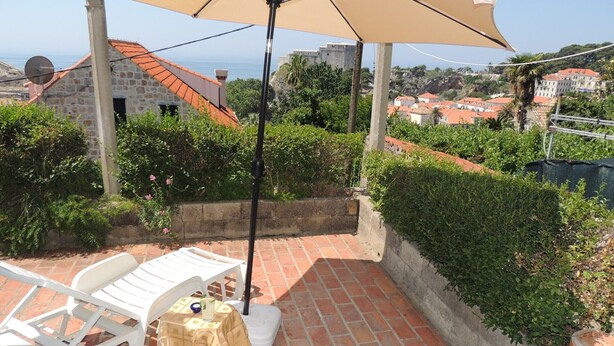 Une terrasse avec des chaises et une table en plastique blanc, un parasol beige, et une vue sur la mer et les bâtiments en arrière-plan.