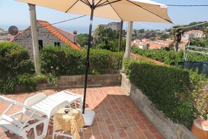 A patio with white plastic chairs and a table, a beige umbrella, and a view of the sea and buildings in the background. - Apartman Sunshine (Dubrovnik)