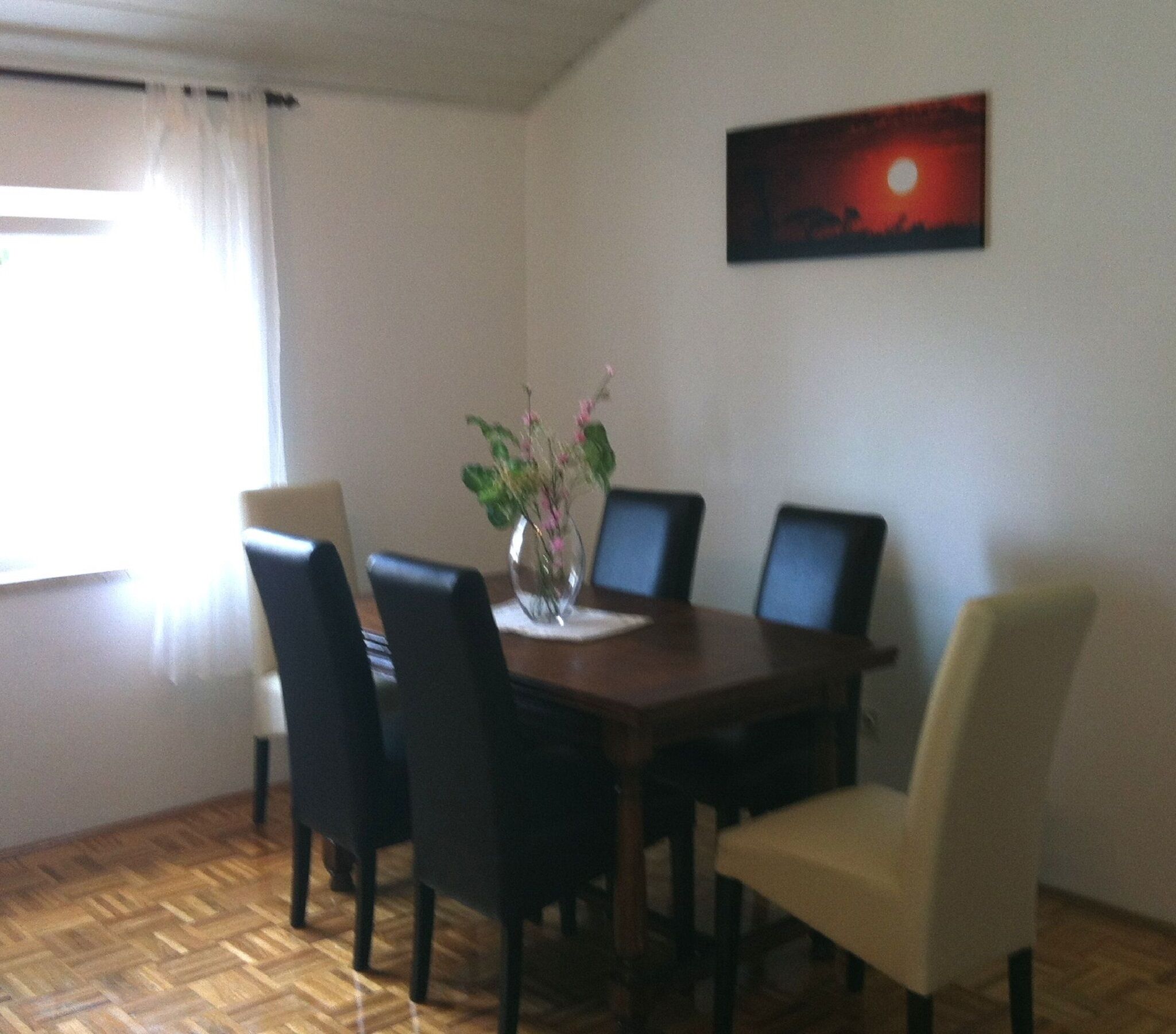 Een eetruimte met een houten tafel, vier stoelen, een vaas met bloemen en een schilderij van een zonsondergang aan de muur.
