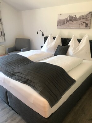 Ein ordentlich bezogenes Bett mit weißer und dunkler Bettwäsche, ein grauer Sessel und eine Wandlampe.