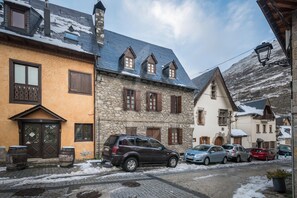 Une rue étroite bordée d’immeubles en pierre, avec des voitures garées et de la neige au sol.