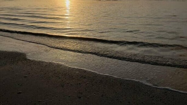 A beach with the sun setting over the horizon, calm waves, and a clear sky.