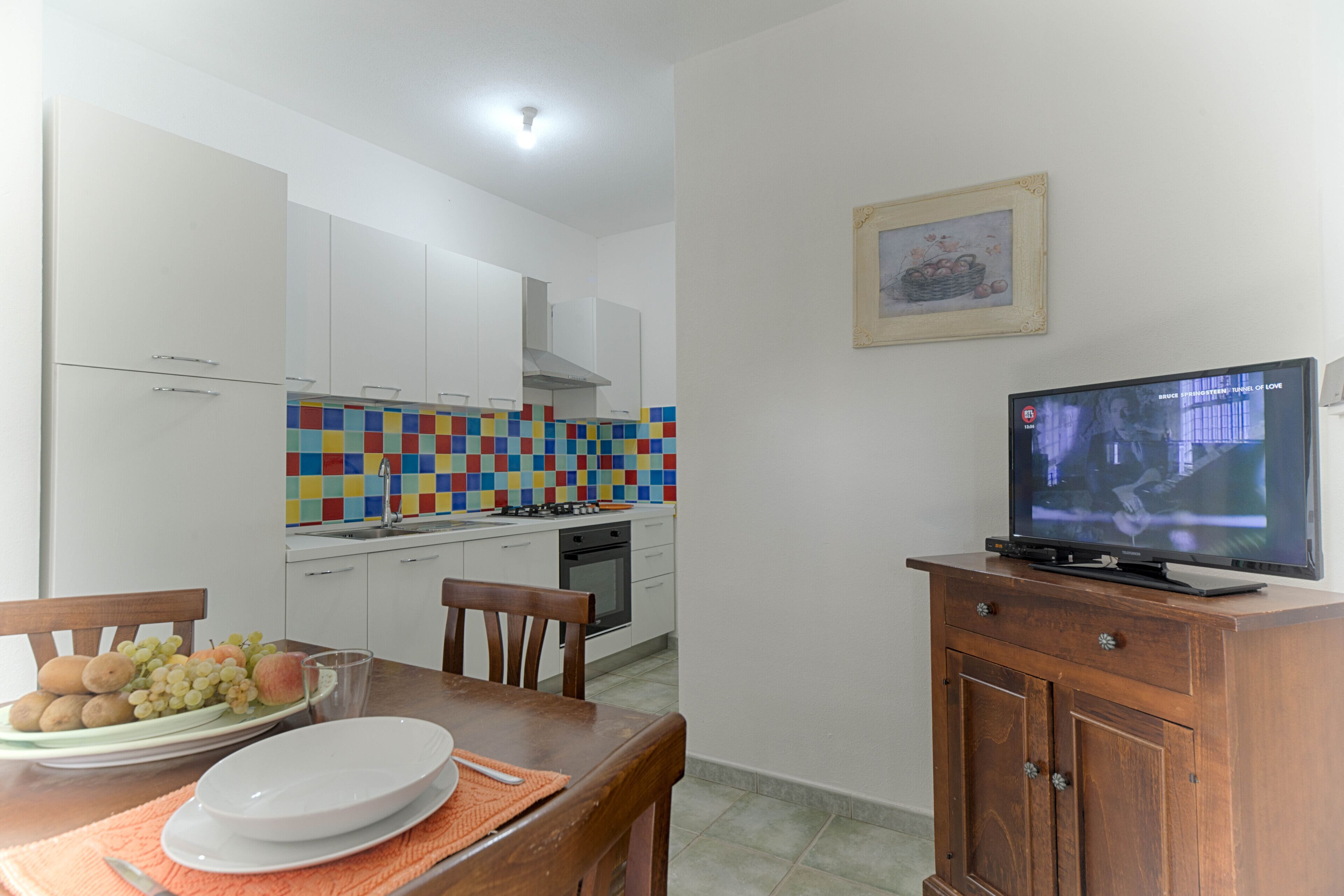 A compact kitchen with white cabinets, a colorful backsplash, and a dining area with a wooden table and chairs. A flat-screen TV is mounted on a wooden cabinet.