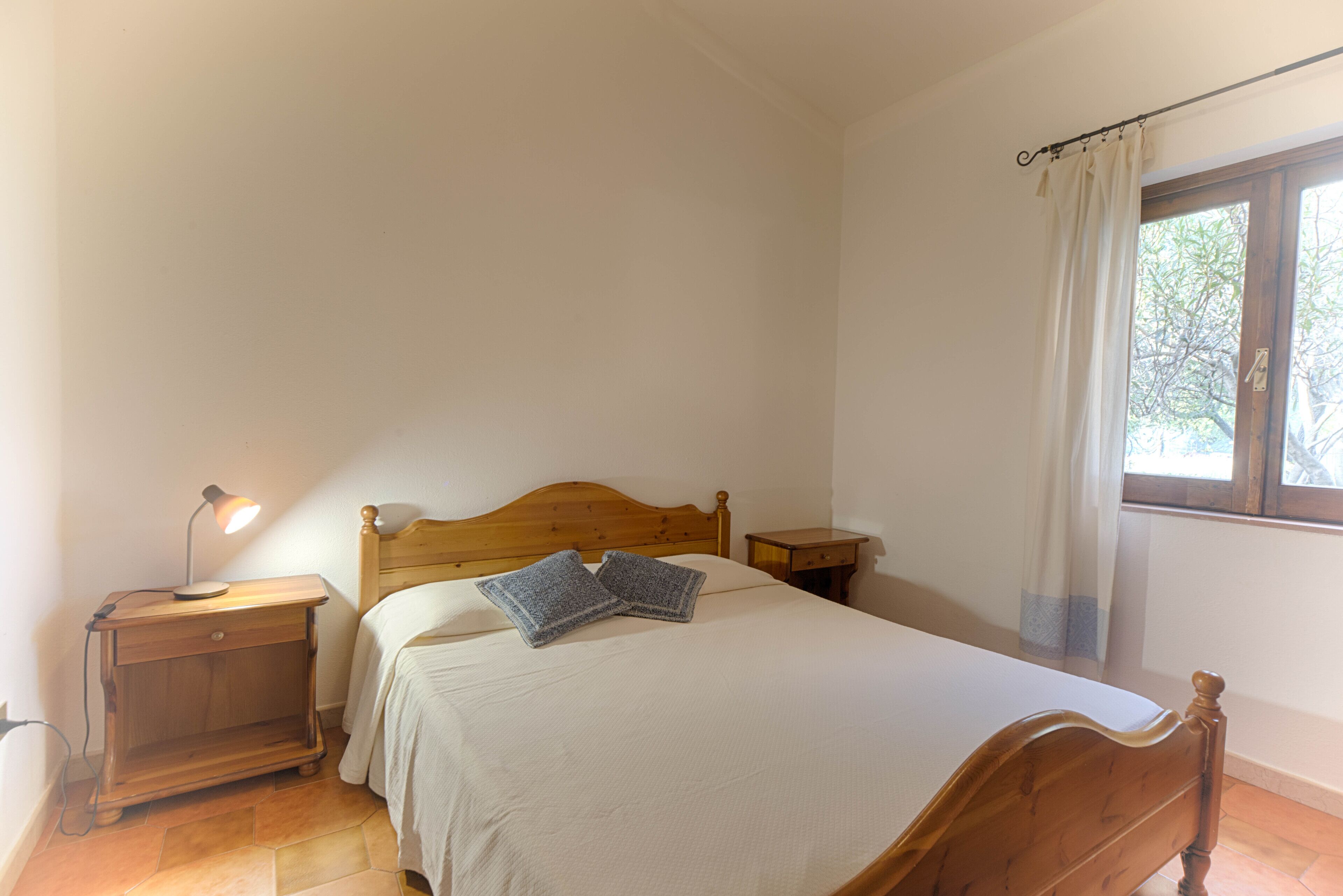 Ein Schlafzimmer mit einem hölzernen Bett, einem Nachttisch mit Lampe und einem Fenster mit Vorhängen.