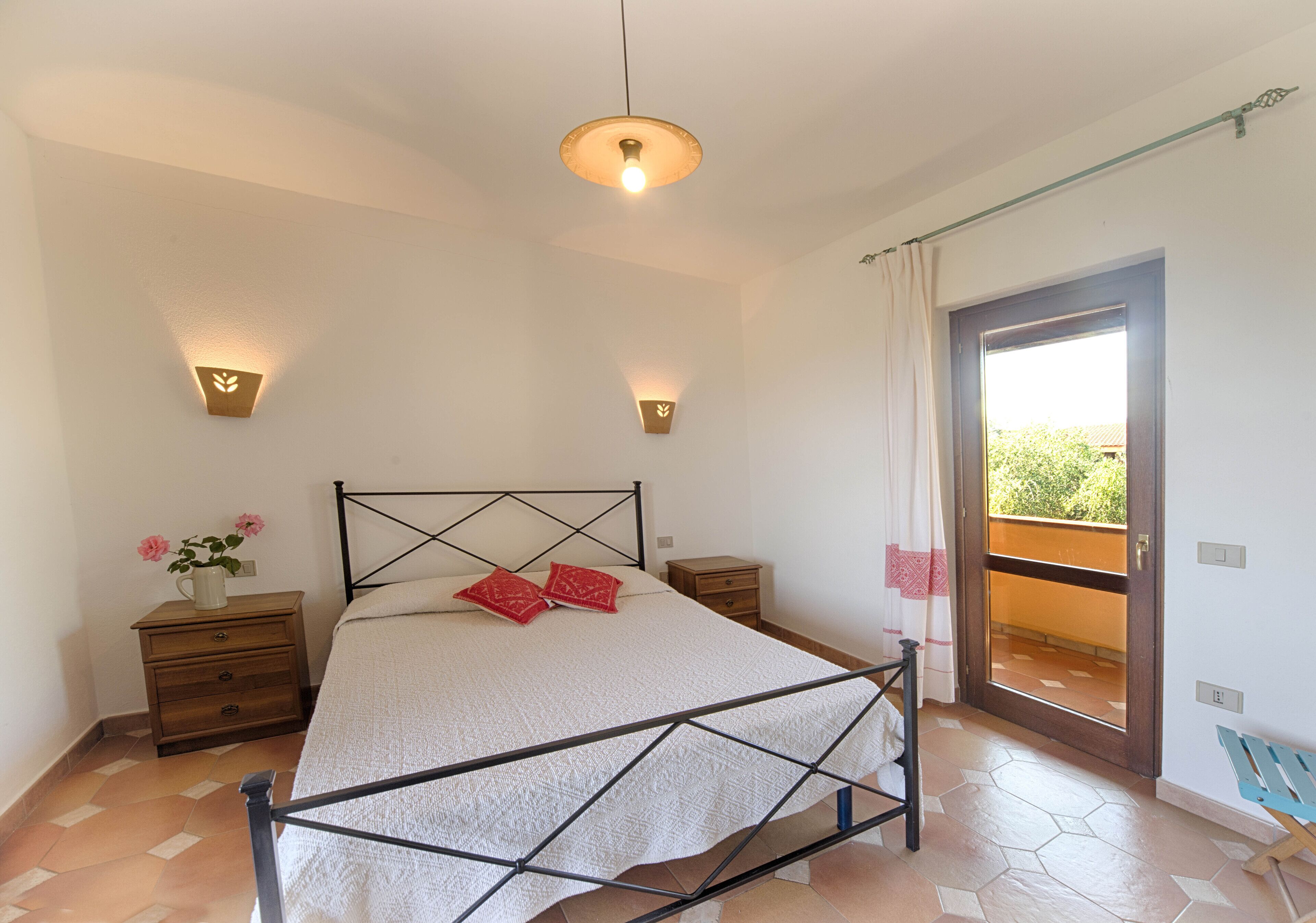 A bedroom with a bed, bedside tables, a nightstand, a wall-mounted light fixture, and a door leading to a balcony.