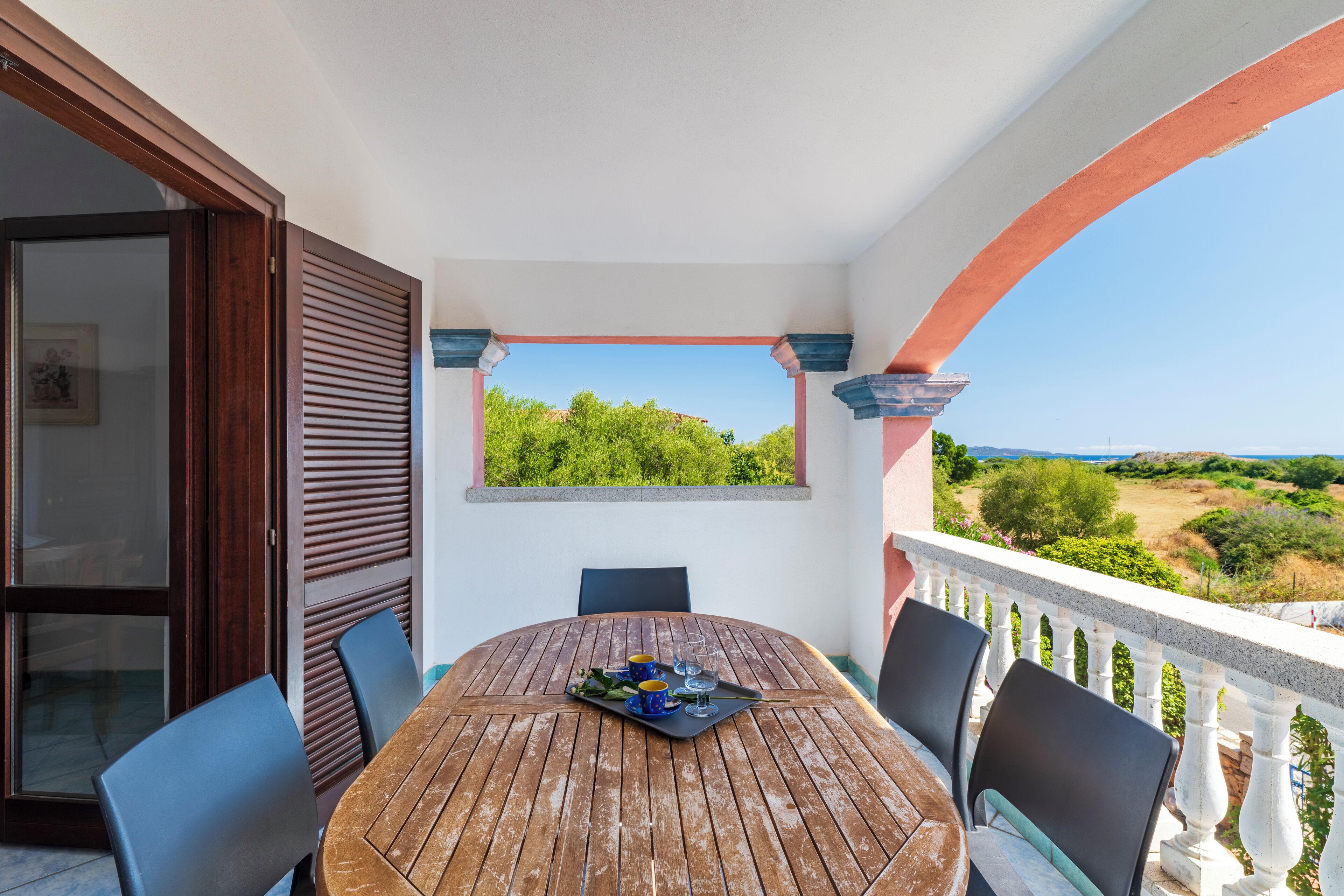 A balcony with a wooden table set for two, chairs, and a view of greenery and hills.