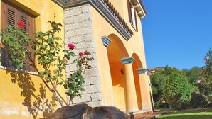 A yellow building with a stone facade, a balcony with wooden shutters, and a garden with a rock feature and a rose bush. - Residence Oasi e Oasi Blu (San Teodoro)