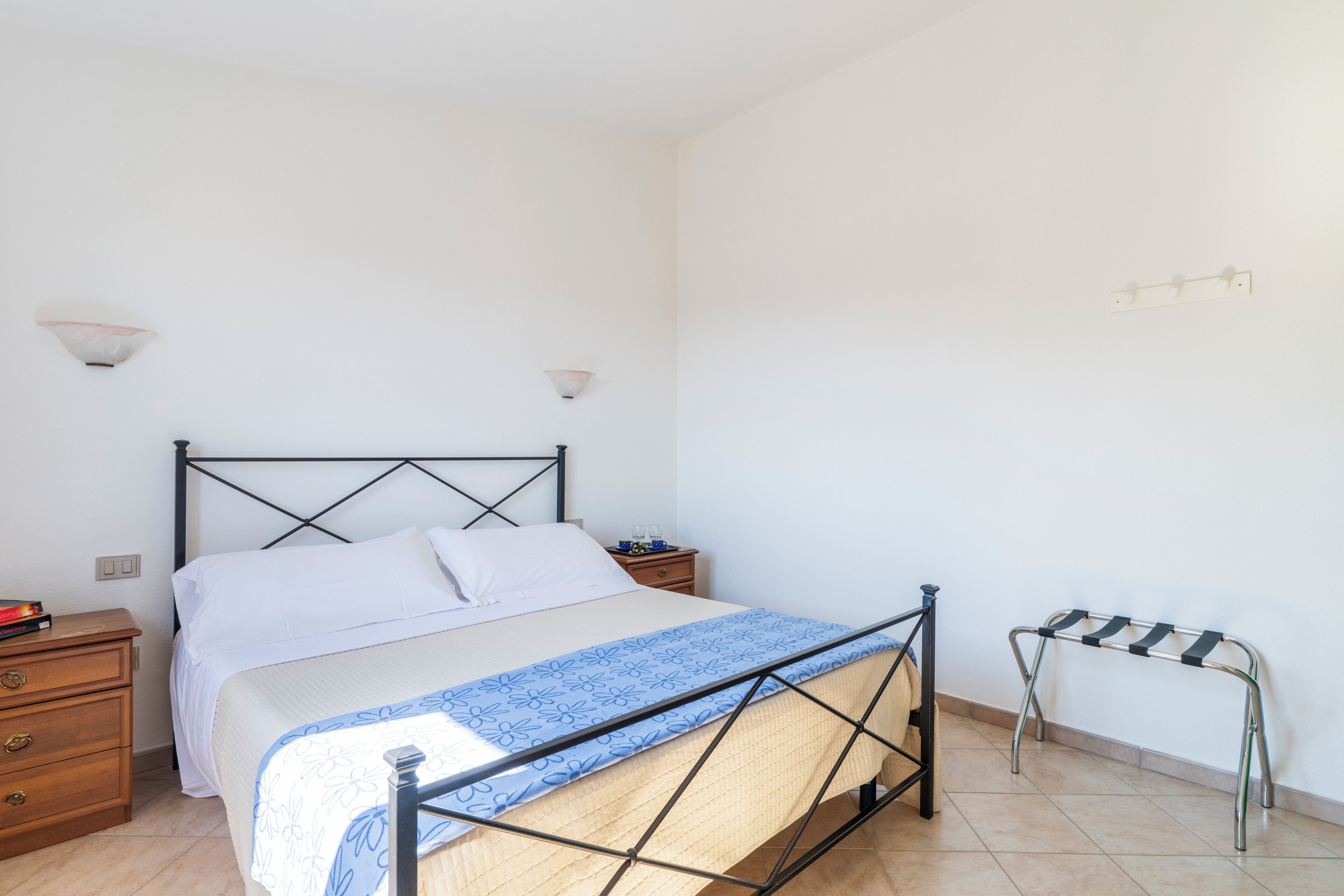 A bedroom with a bed, bedside tables, a nightstand, a wall-mounted light fixture, and a metal rack.