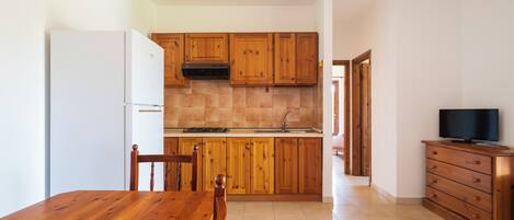 Una cocina con armarios de madera, un refrigerador blanco, una mesa de comedor con sillas y un televisor sobre un mueble de madera.