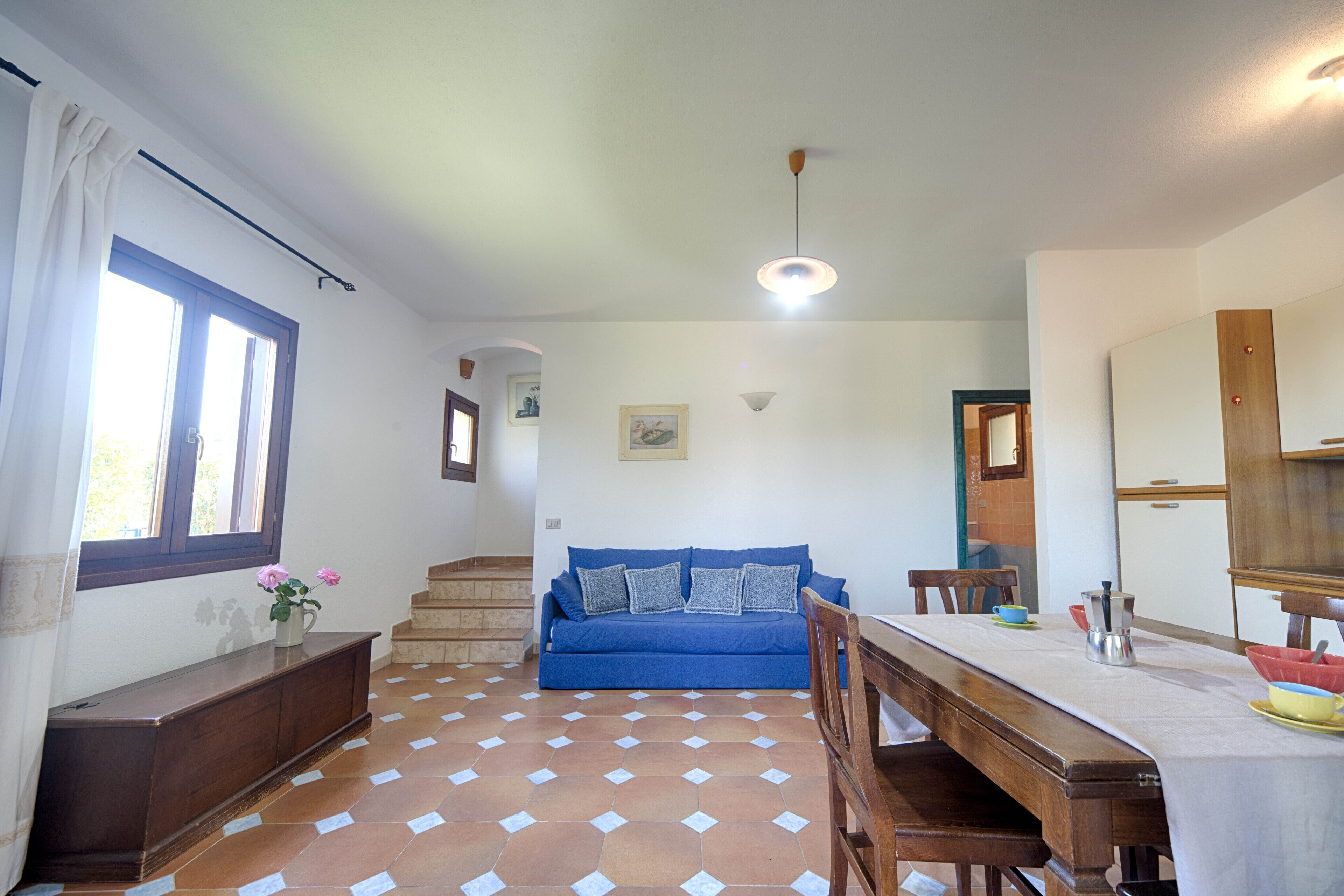 A spacious living area with a blue sofa, wooden dining table, and a tiled floor.
