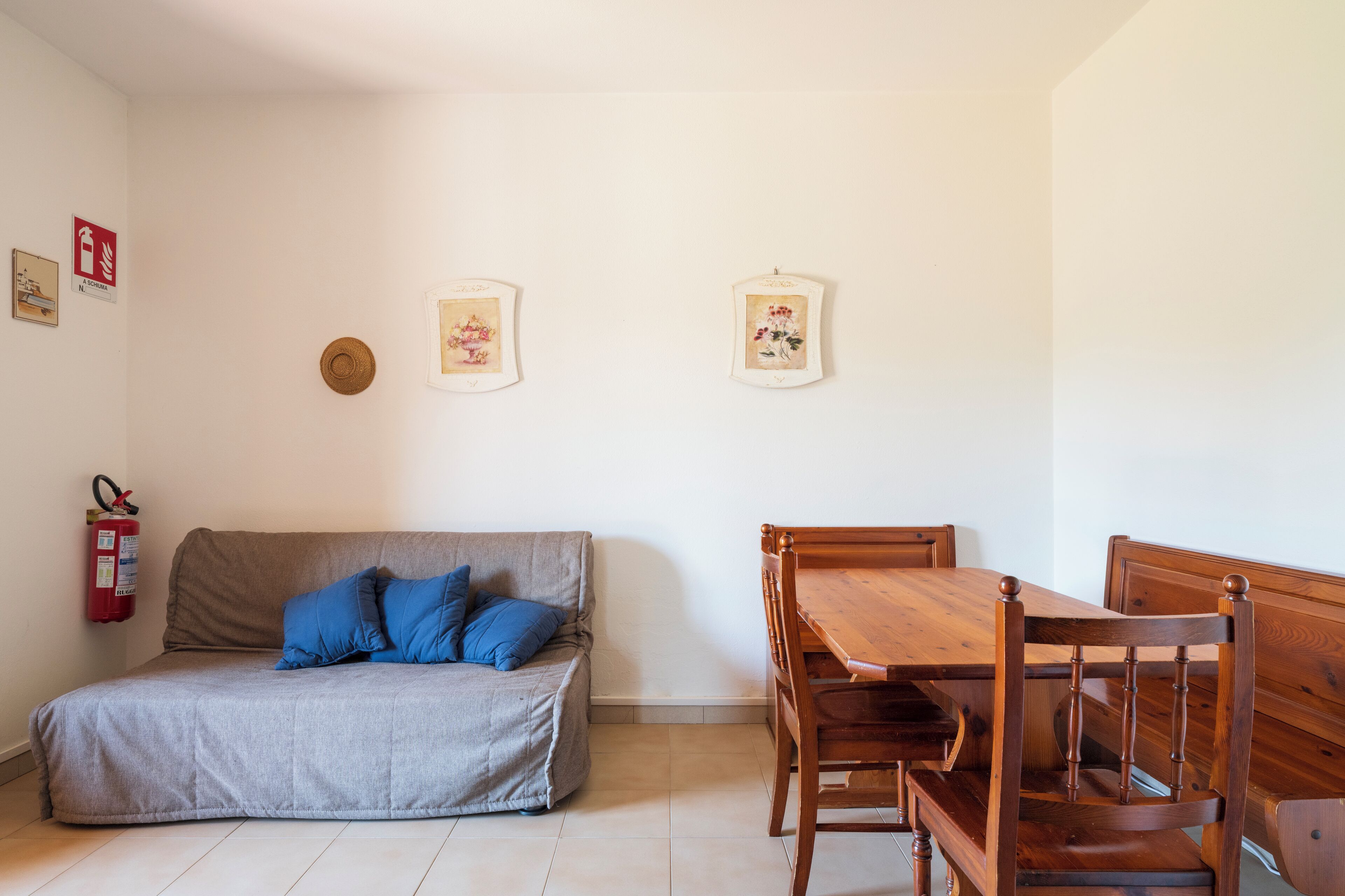 A room with a sofa, a wooden dining table, and chairs. There is a fire extinguisher on the wall.