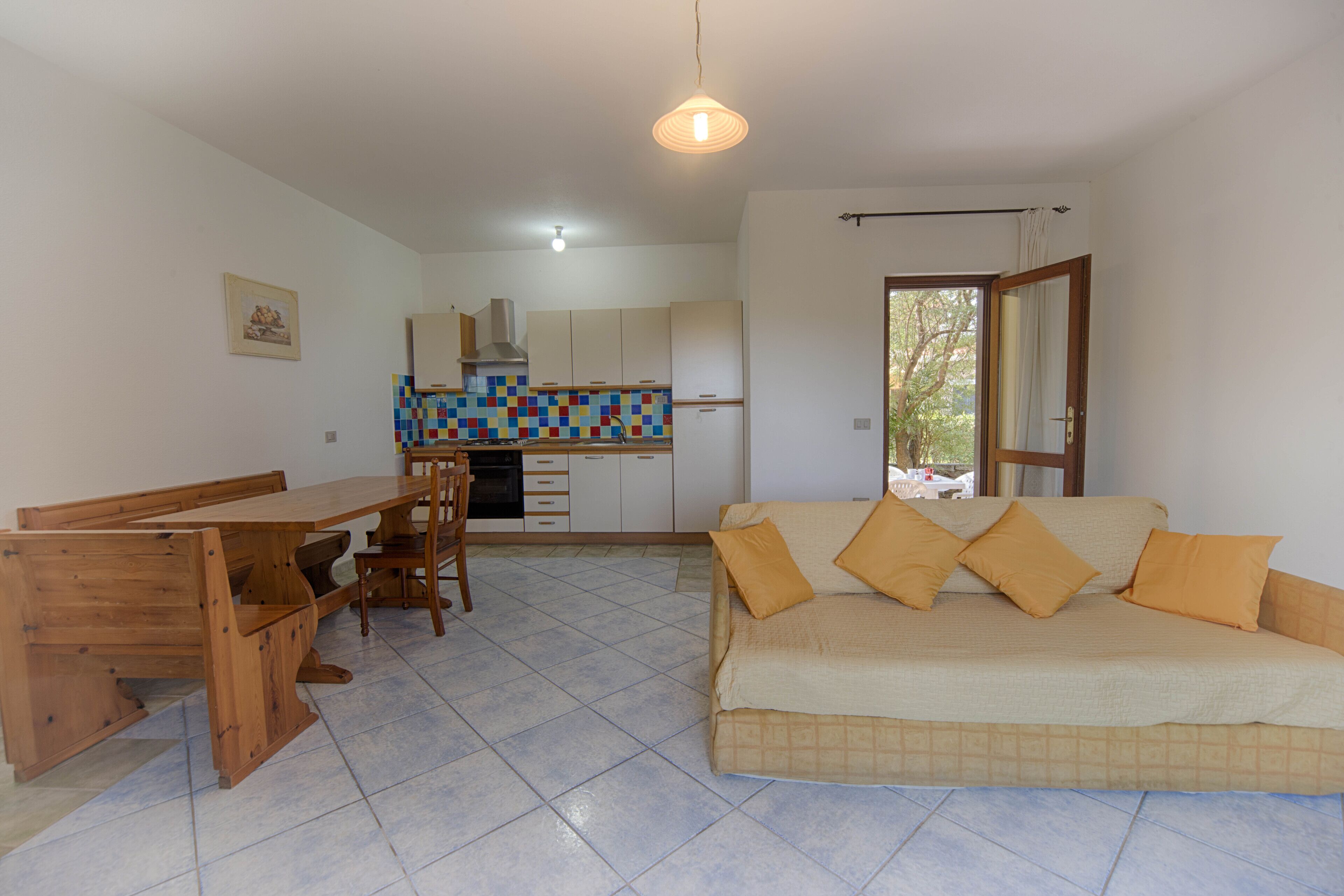 Amplia sala de estar con sofá, mesa de comedor de madera y sillas. Se ve una cocina con armarios y salpicadero de azulejos al fondo.