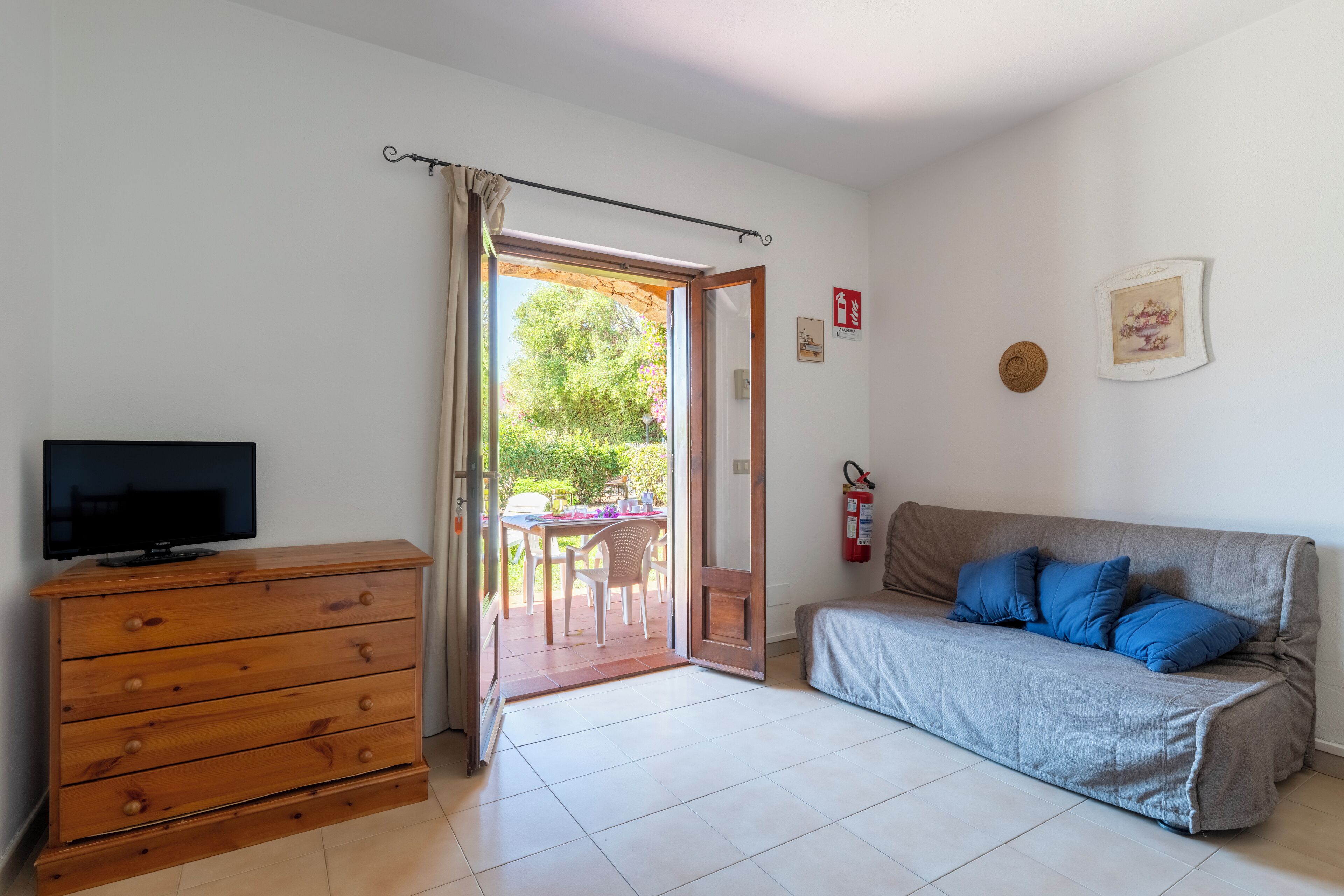 Una stanza con una cassettiera in legno, un divano grigio con cuscini blu, una televisione sulla cassettiera e una porta che dà su un'area esterna con tavolo e sedie.