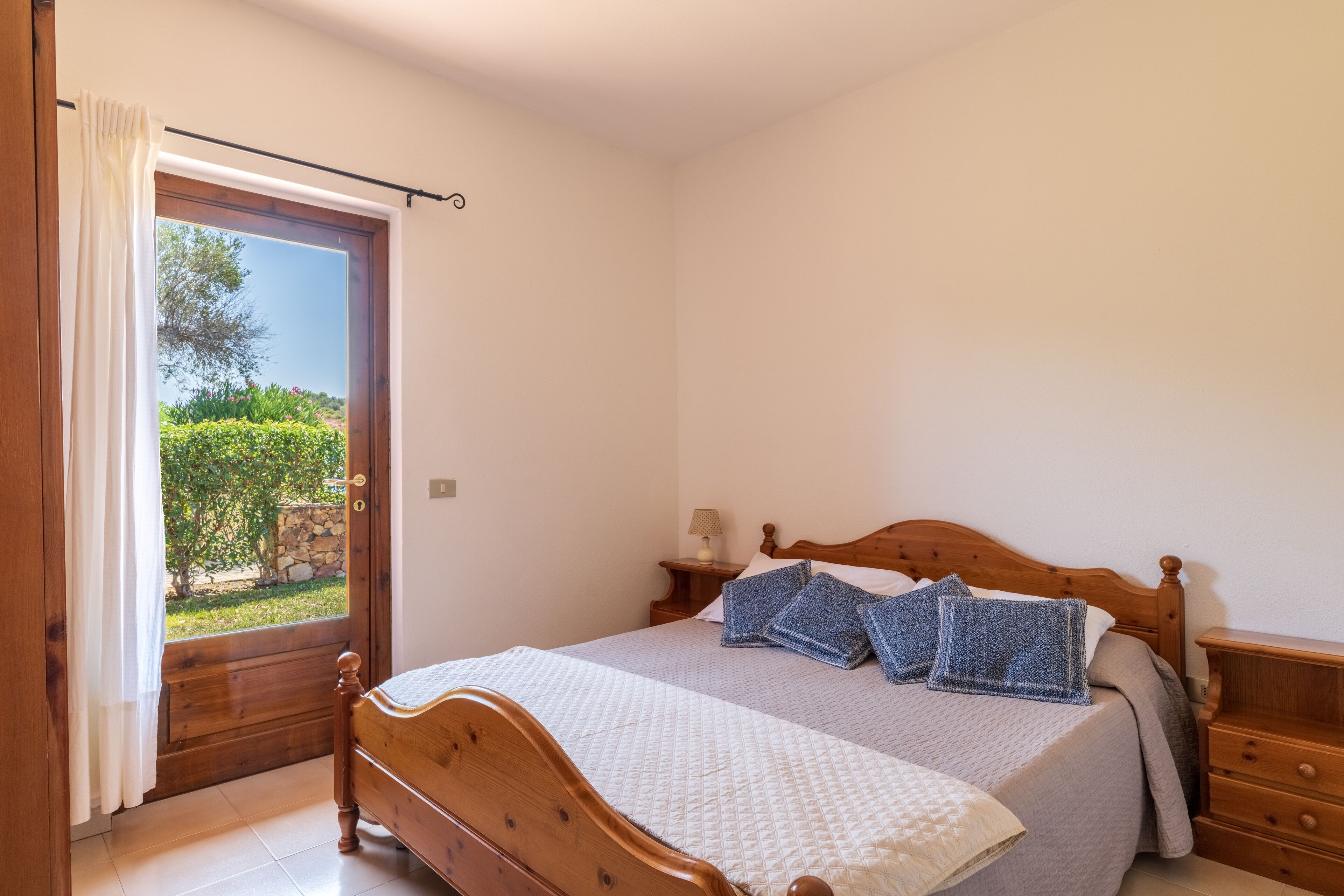 A bedroom with a wooden bed, a nightstand, a lamp, and a view of greenery through a door.
