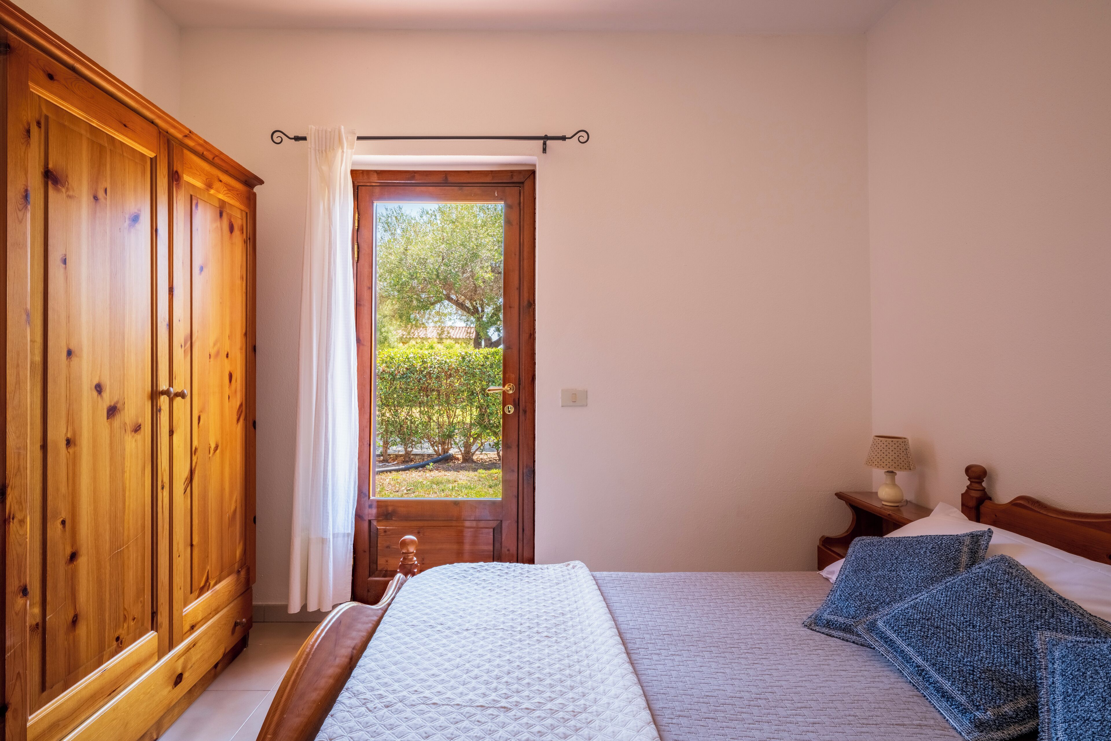 A bedroom with a large wooden wardrobe, a bed with blue pillows, a nightstand with a lamp, and a view of a garden through an open door.