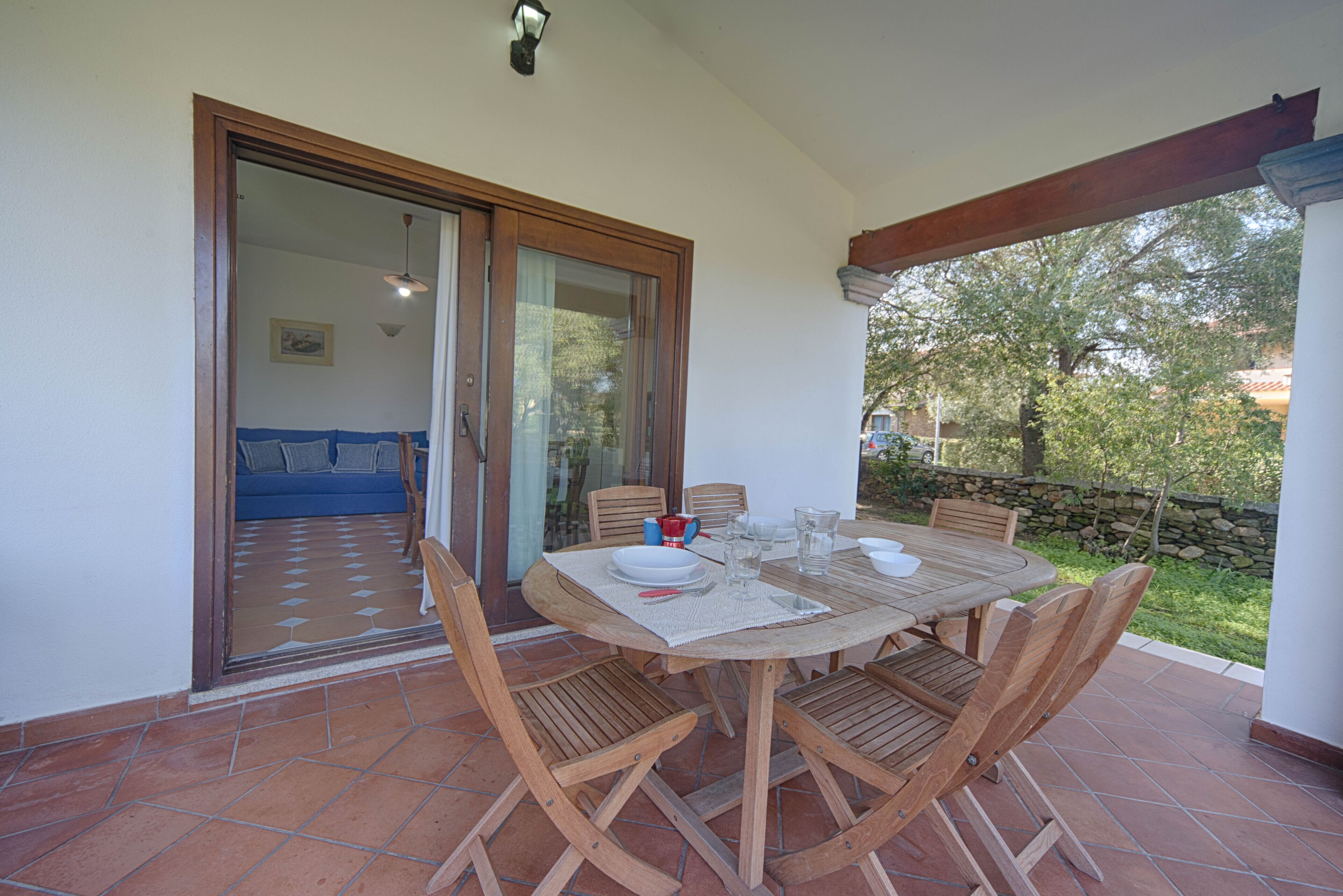 A patio with a round wooden table and chairs, a glass door leading to an interior room, and a stone wall in the background.